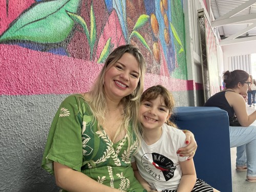 Mulher sorrindo sentada ao lado de uma menina, ambas com expressões alegres, em frente a uma parede colorida com arte mural.