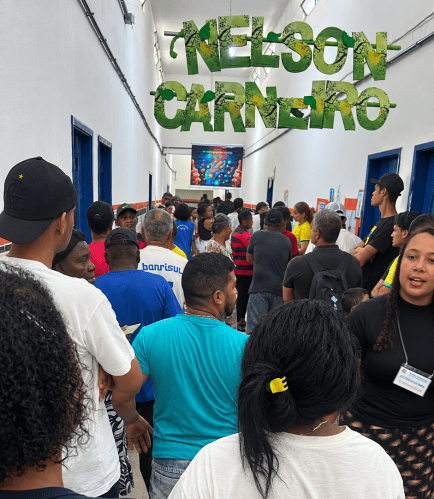 Fila de pessoas em um corredor, com paredes brancas e portas azuis, em um evento com o nome 'NELSON CARNEIRO' destacado no topo.