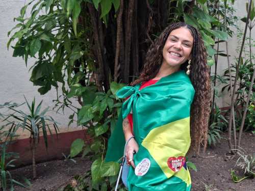 Mulher sorridente vestindo uma capa com as cores da bandeira do Brasil, posando em frente a uma planta verde.